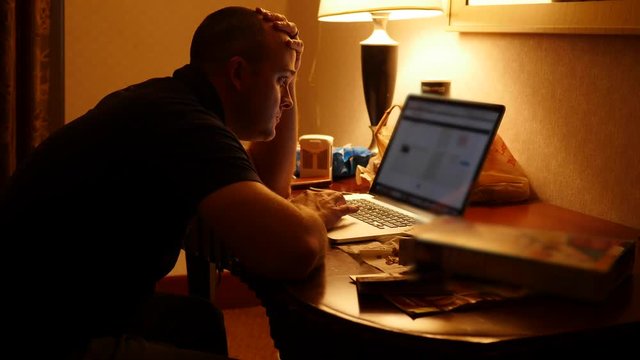 A man working in dark hotel room at night on computer gets up