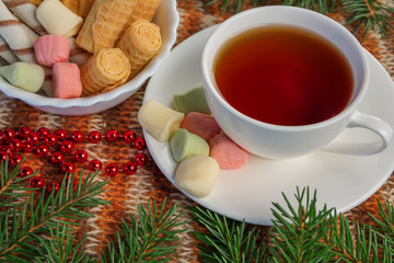The Christmas tea. A Cup of tea. Cookies, marshmallows and marmalade on a saucer. Christmas decor. Red beads. Spruce branches. In warm tones. Knitted background. Studio light.