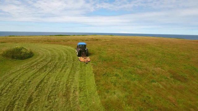 Aerial rotating shot farmer cuts the grass on field by ocean cove