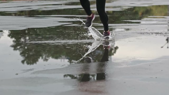 Closeup shot of fit female legs in sport tights and sneakers running through puddles in slow motion and slapping against water on pavement