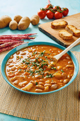 Bean soup in blue dish and wooden spoon surrounded by potatoes, tomatoes, beans in pod, croutons on blue wooden table