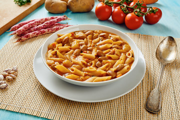 Pasta with beans in white dish, potatoes, carrots, tomatoes in background