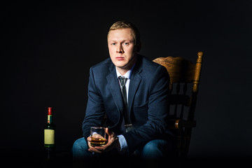 Businessman wearing a jacket with  glass of whiskey sitting on  chair. Bottle alcohol at dark studio background. Concept  choice, privileges, solution problems