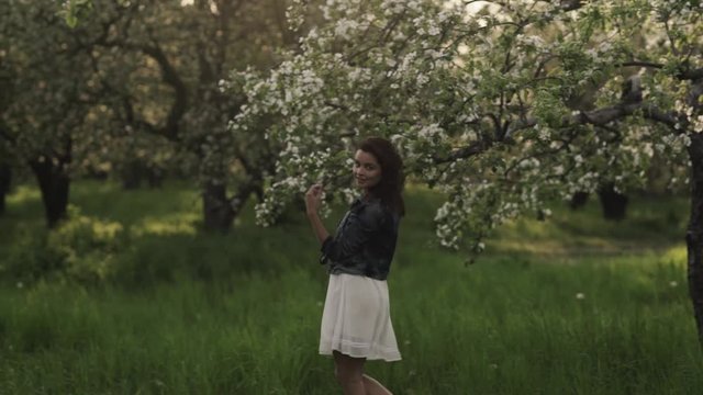 Young pretty smiling  caucasian woman in jeans jacket and white dress walks in a blooming spring garden. Youth, freshness, beauty, happiness concept.