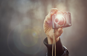 Adult man hand holding a compact digital point-and-shoot photo camera over out of focus outdoor background. Taking outdoor pictures with a digital compact camera. Lens flare and copy space.