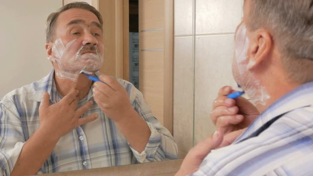 Senior man of shaving in the bathroom. It uses shaving foam and disposable razor. The concept of self-care
