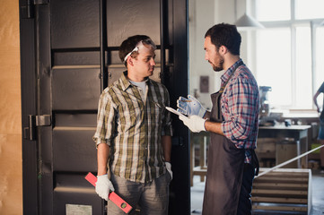Two carpenters are talking while holding papers and ruler. One of them is wearing safety mask. Carpentry shop background