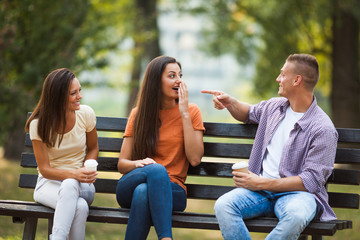 Three friends are sitting on bench in park and talking.