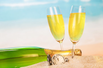 Bottle, two glasses with champagne, clock in sand against sea.