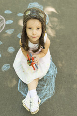 Little girl wearing a pilot hat with goggles holding in hand biplane toy sitting down on asphalt after drawing clouds with chalk. Concept of child imagination. Little child girl plays astronaut © beatricemihaela