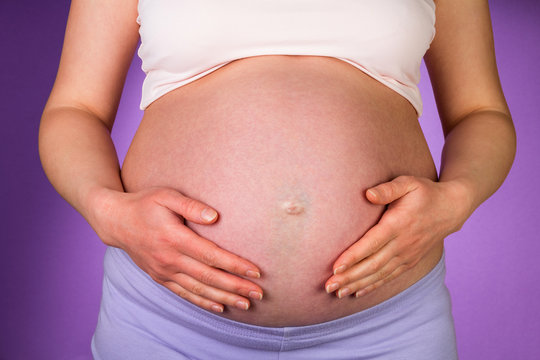Pregnant woman embracing belly her hands on purple background.
