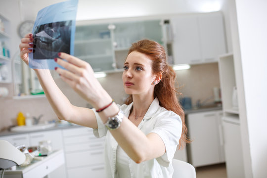  A red-haired female dentist carefully examines an X-ray image of a patient's teeth, ensuring precise diagnoses and comprehensive dental care.