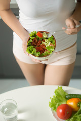 Female wearing underwear panties and white color tank top holding a bowl of green salad against her waist near the kitchen table, preparing to it with a fork, close up. Vertical, weight loss concept