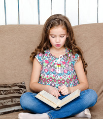 Cute little girl is reading the book on the sofa