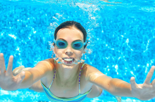 Child swims in pool underwater, funny happy girl in goggles has fun under water and makes bubbles, kid sport on family vacation
