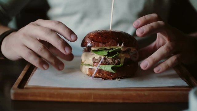Tasty burger is great on a wooden tray. A man takes his hands and eats, try the taste