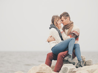 Young attractive couple in knitted sweaters on a cliff on sea shore, hugging and holding hands, creative toning