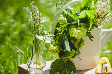 Gießkanne mit Maiglöckchen in einer Vase stehen auf dem Hocker im Garten. 