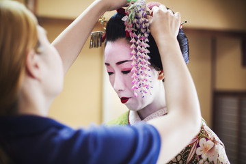 A geisha or maiko with a hair and make up artist creating the traditional hair style and make up. An elaborate flower decoration with long tassels being fixed in her hair. 
