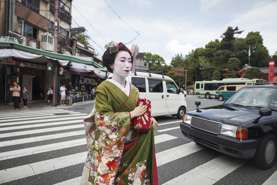 A woman dressed in the traditional geisha style, wearing a kimono and obi, with an elaborate hairstyle and floral hair clips, with white face makeup with bright red lips and dark eyes crossing a street. 