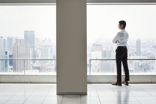 A businessman in the office standing by a large window with arms folded, looking over the city.
