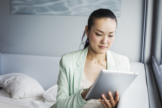A business woman preparing for work, sitting on a bed using a digital tablet.