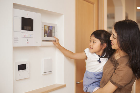 Family home. A woman and her daughter looking at a screen on the wall, controls of an entry phone or doorway.