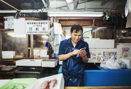 A traditional fresh fish market in Tokyo. A man in a blue apron standing behind the counter of his stall, using a smart phone covered with protective plastic. 