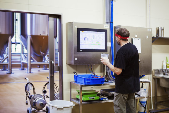 A brewer with a red bandana checking a computer monitor and testing the brewed beer. 