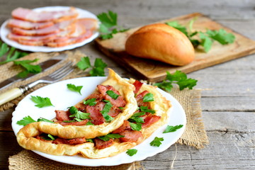 Homemade bacon omelette. Delicious omelette stuffed with bacon and parsley on a plate, bacon slices on a plate, bread roll, fork, knife, cutting board, fresh parsley sprigs on old wooden background