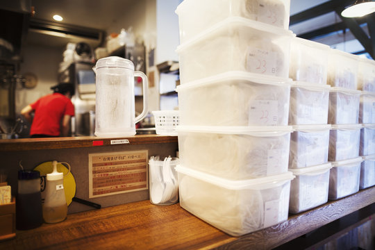 The ramen noodle shop.  Staff preparing food in a kitchen, and rows of plastic boxes of food. 