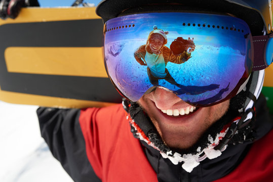 Happy snowboarder in mask with friend taking shot in reflection
