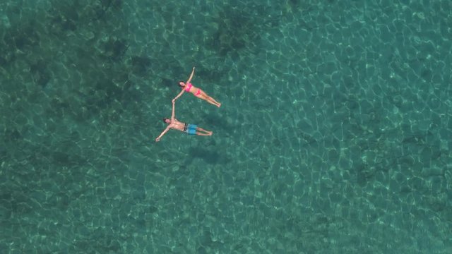 AERIAL: Happy couple swimming starfish float holding hands in crystal clear sea