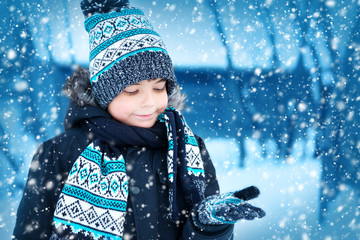 Boy in knitted hat, gloves and scarf outdoors at snowfall. Child in winter clothes