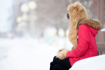 Portrait of unhappy beautiful young woman wearing red winter coat and cute warm fur hat and mittens sitting alone on the bench outdoors, waiting for somebody or feeling cold, copyspace