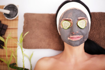 Beautiful young woman is getting facial clay mask at spa, lying with cucumbers on eyes