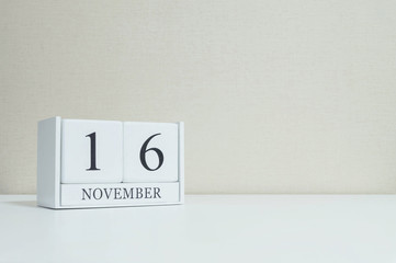 Closeup white wooden calendar with black 16 november word on blurred white wood desk and cream color wallpaper in room textured background with copy space , selective focus at the calendar