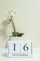 Closeup white wooden calendar with black 16 november word with white orchid flower on white wood desk and cream color wallpaper in room textured background , selective focus at the calendar