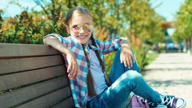 Cute schoolgirl 7-8 years in sunglasses sitting on the bench and smiling at camera in the park. Zooming