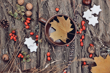 Top view of a mug of tea covered with an autumnal maple leaf, surrounded by walnuts, rose hips, on a wood barkw