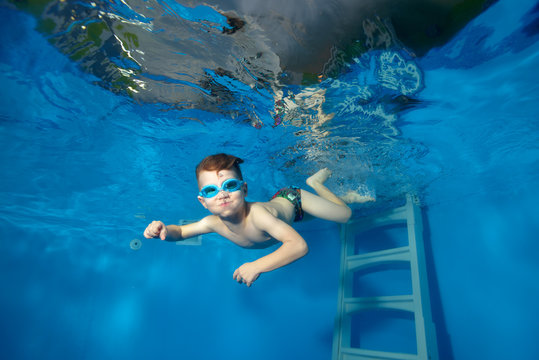 Little boy swims underwater in the pool, looking at me and smiling on a blue background. The view from under the water. Portrait. Horizontal orientation