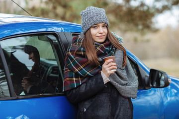 Outdoor portrait of young caucasian woman, holding a cup of takeaway coffee in forest park on cold season day. Dressed in an elegant leather jacket, grey hat and big cozy scarf