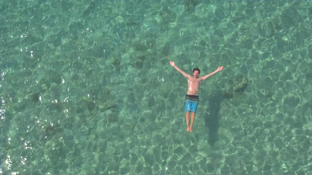 AERIAL: Young man swimming starfish float and relaxing in crystal clear ocean