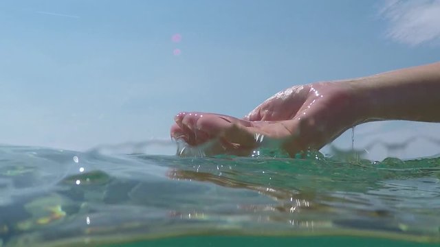 SLOW MOTION SPLIT UNDERWATER: Hand scooping water, drops falling on sea surface