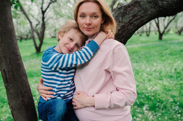 spring portrait of happy pregnant mother enjoying warm day with child son in blooming apple garden