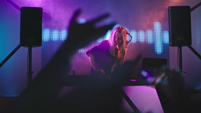 Lockdown of blond female DJ dancing behind mixer at party in nightclub 