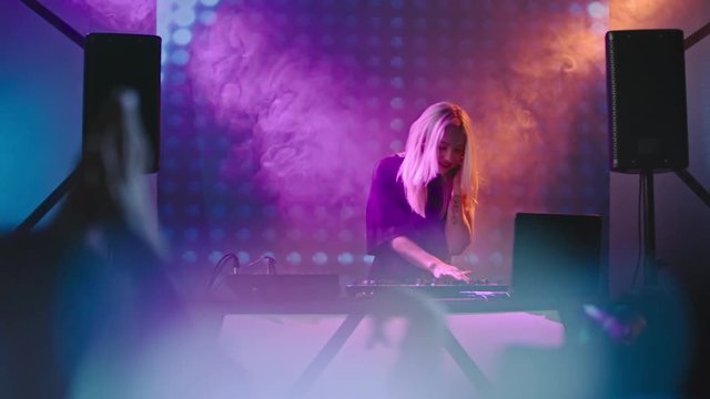 Blond female DJ dancing behind mixer console surrounded by smoke and colorful LED projection