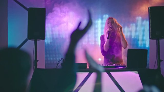 Blond female DJ dancing behind decks, then picking up microphone and singing into it for cheering crowd