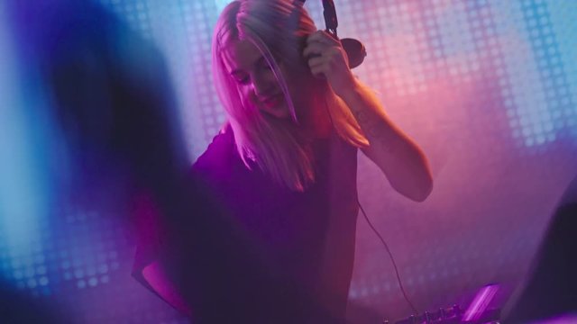 Dutch angle of seductive blond female DJ dancing behind decks in nightclub 