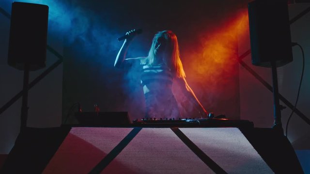 Female DJ with blond hair jumping and singing behind decks on concert in nightclub
